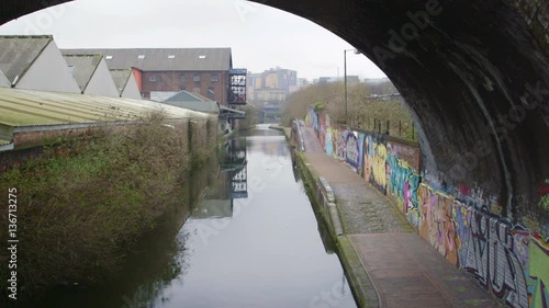Along a graffiti sprayed canal walkway, underneath a blackened railway arch, in the heart of industrial urban Birmingham the reflections in the water show factories and bushes.