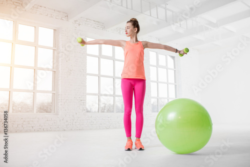 Young woman in colorful sportswear lifting dumbbels in the white gym with green fitness ball on the floor © rh2010