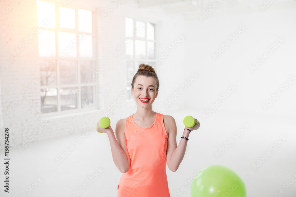 Portrait of young woman in sportswear with dumbbells and fitness ball in the white gym Portrait of young woman in sportswear with dumbbells and fitness ball in the white gym