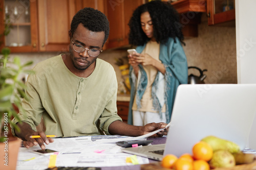 Busy serious African male using cell phone while calculating family expenses and doing paperwork, sitting at kitchen table in front of laptop computer, his wife standing in background with gadget