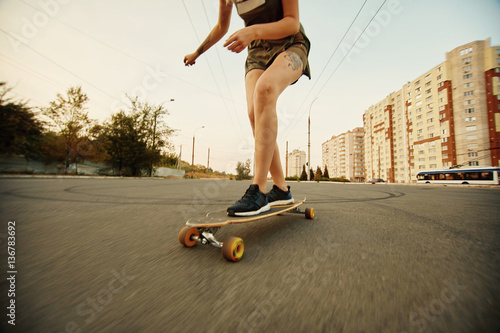 Beautiful young girl with tattoos riding on his longboard on the road in the city in sunny weather. Extreme sports. Rear view of motion photo