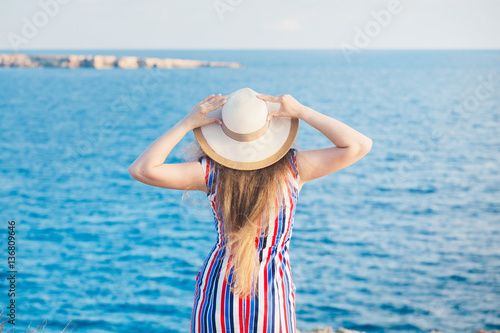 Back view of woman standing in summer dress and hat looking out towards blue ocean and sky