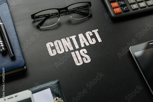 Black chalkboard with business accessories (notepad, magnifying glass, fountain pen, tablet, phone, glasses and calculator) and text CONTACT US. Top view.