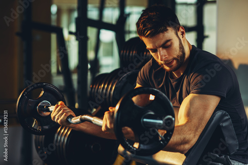 Handsome man doing biceps lifting barbell on bench in a gym