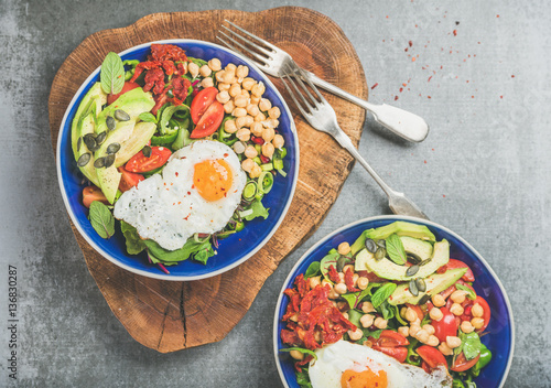 Healthy breakfast bowls with fried egg, chickpea sprouts, seeds, vegetables and greens over grey concrete background, top view. Clean eating, dieting, healthy lifestyle, detox, vegetarian food concept