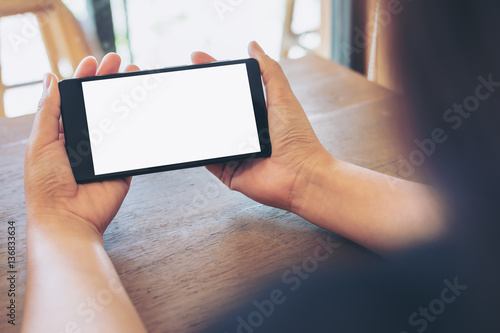Mockup image of hands holding black mobile phone with blank white screen on vintage wood table in cafe