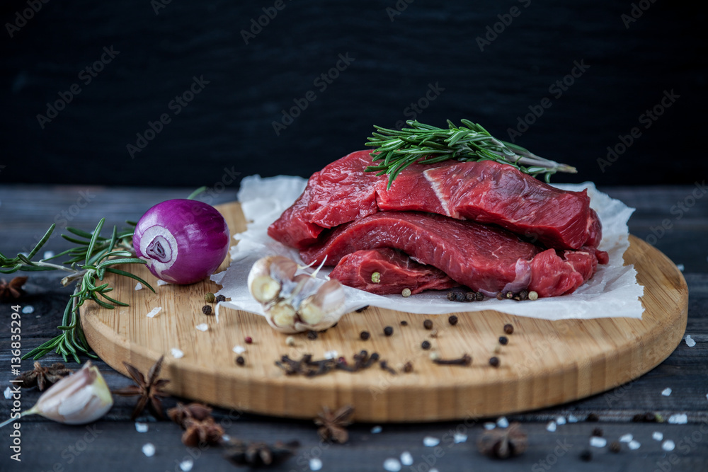 Raw meat beef steaks slices ready for roasting on a wooden cutting board with fresh organic vegetables, onion, garlic, spices, peppercorns, rosemary, sea salt and herbs Raw meat beef steaks slices ready for roasting on a wooden cutting board with fresh organic vegetables, onion, garlic, spices, peppercorns, rosemary, sea salt and herbs