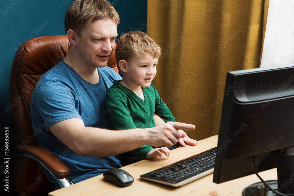 Father and little son using computer at home. The father with the son watch video and cheerfully spend time together. Stady in the computer. Father and little son using computer at home. The father with the son watch video and cheerfully spend time together. Stady in the computer.