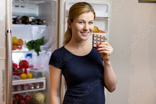 Healthy Eating Concept. Happy woman with chocolate bar standing at the opened fridge with fruits, vegetables and healthy food