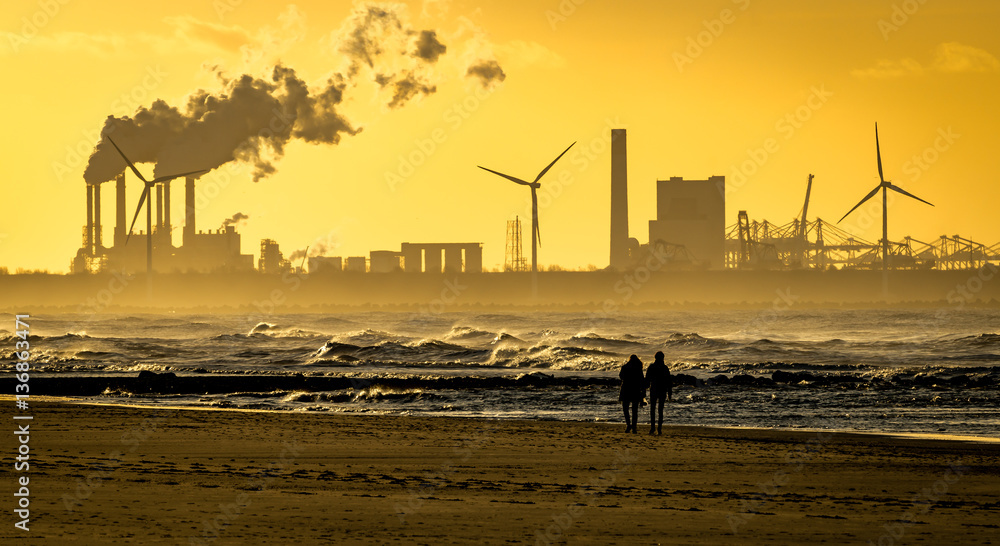 Walking couple on beach with smoke stacks and wind turbines on horizon in sunset