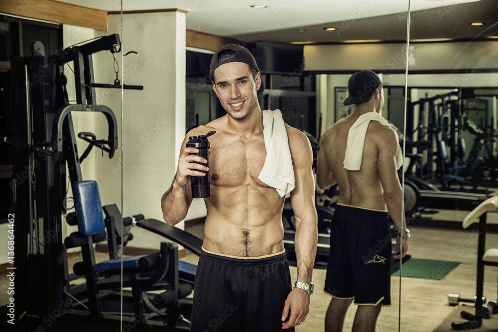 Fototapeta premium Attractive athletic shirtless young man holding protein shake in a blender in gym while looking at camera