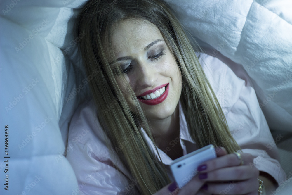 Young beautiful woman smiling under the bed covers. Relaxing at home. Woman laying in bed and texting. Young beautiful woman smiling under the bed covers. Relaxing at home. Woman laying in bed and texting.