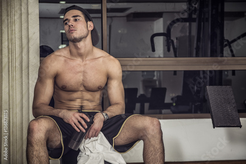 Muscular, shirtless young man resting in gym during workout, showing muscular torso, pecs and abs, holding a towel in his hands, sitting on bench