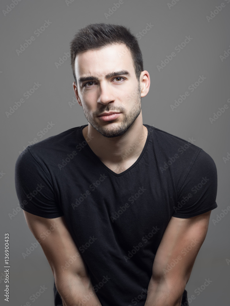 Moody portrait of fit muscular serious man wearing blank black t-shirt looking at camera suspiciously over gray studio background. 