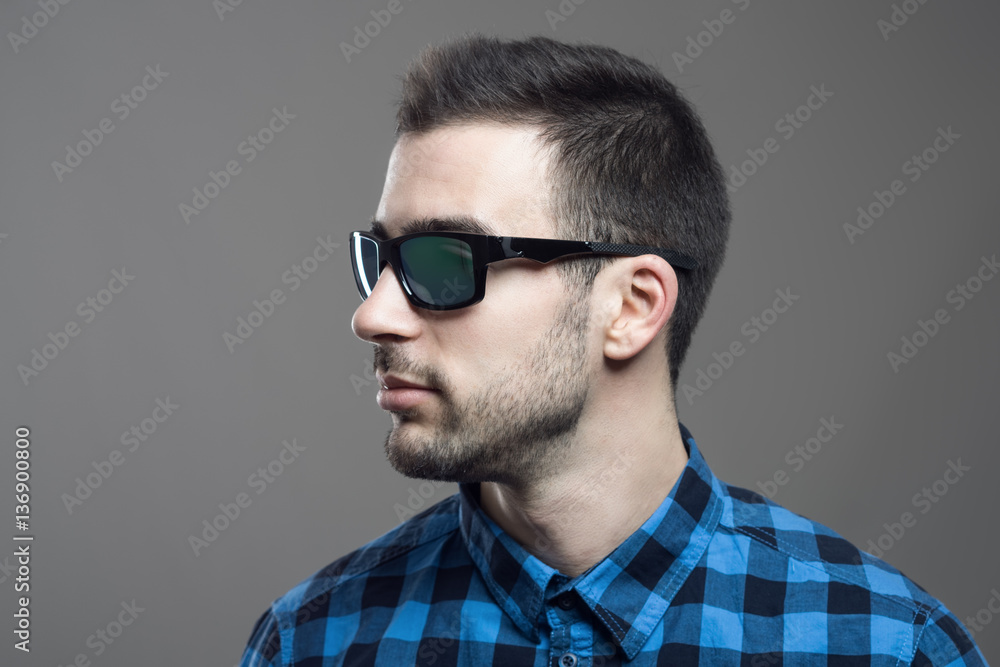 Profile view of young bearded man in plaid shirt wearing sunglasses looking away over gray studio background. Profile view of young bearded man in plaid shirt wearing sunglasses looking away over gray studio background.
