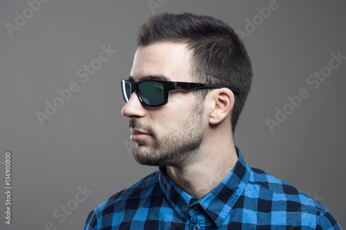 Profile view of young bearded man in plaid shirt wearing sunglasses looking away over gray studio background. 