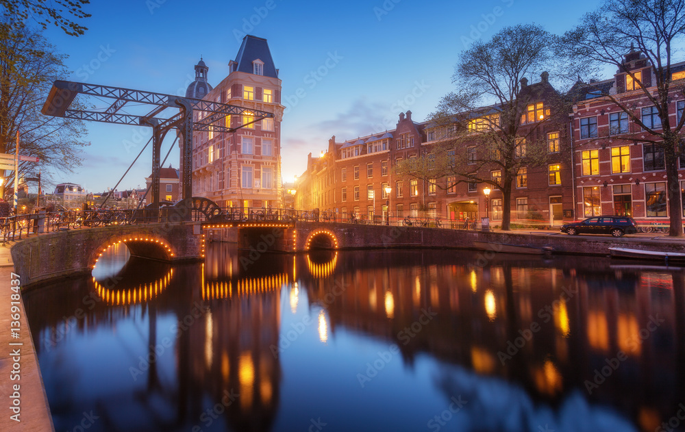 Colorful cityscape at sunset in Amsterdam, Netherlands. Reflected city lights in water with sunlight and blue sky at twilight. Night illumination of buildings. Travel. Beautiful street with bridge