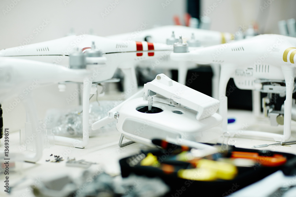 Several high-tech drones standing on white table in modern workshop with controller and assembling tools in front