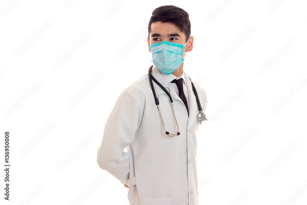 beautiful young brunette male doctor with stethoscope in uniform and mask posing isolated on white background beautiful young brunette male doctor with stethoscope in uniform and mask posing isolated on white background