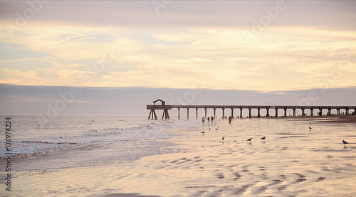 Ocean pier jetty at spectacular sunset. People walking on beach. Peaceful scene, calming waves, pastel cloudy sky, coast. Soft light