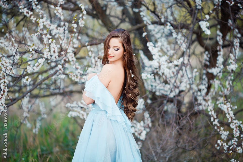 Spring touch. Happy beautiful young woman in blue dress enjoy fresh flowers and sun light in blossom park at sunset showing her naked back and shoulder.