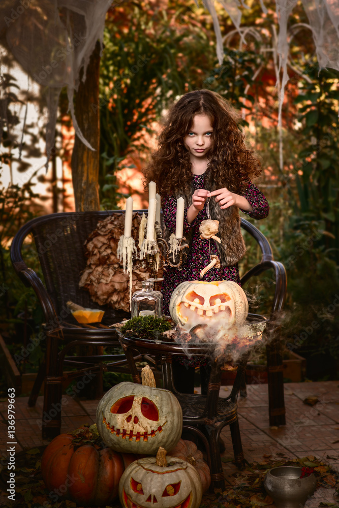 Happy Halloween Concept! Cute little witch cooking a poition in the decorated garden.  Beautiful child girl in witch costume  with halloween decoration:  book of spells, poition and big pumpkin.