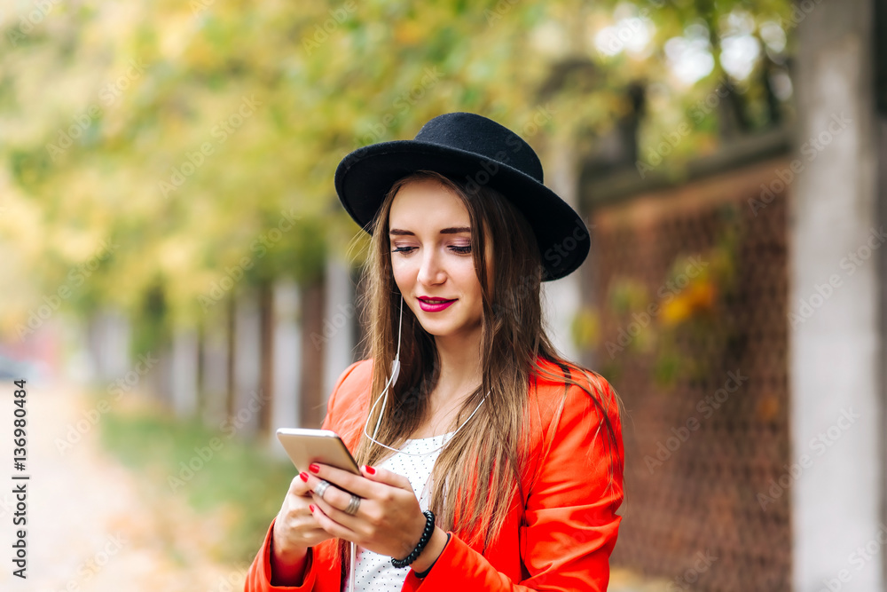 young stylish pretty woman with black hat in red jacket posing in the city streets. Girl holding a phone and writing a text message