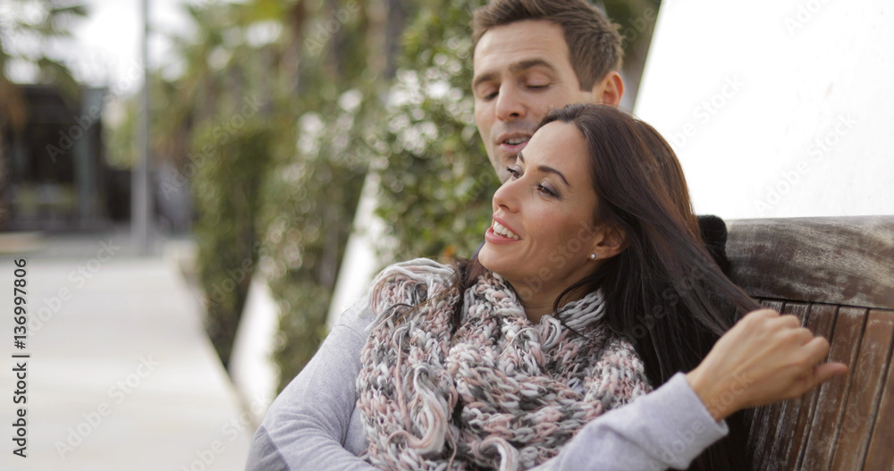 Trendy attractive young woman relaxing with her beau as they sit together on a wooden park bench looking into the distance with a smile focus to her. Trendy attractive young woman relaxing with her beau as they sit together on a wooden park bench looking into the distance with a smile focus to her.