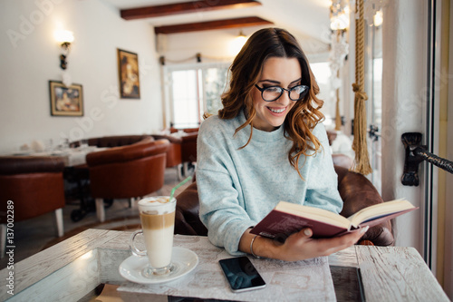 Fotografi Young  beautiful woman in glasses reading interesting book in cafe