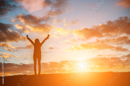 Konstfotografi Silhouette woman at sunset standing elated with arms raised up.