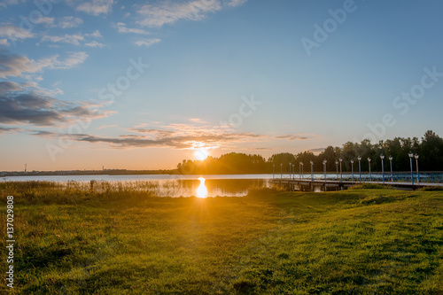 Evening lights of orange sunset over lake in a Valday region village at early spring, Russia. Outdoor nature landscape with sun goes down the lake and lights reflection in calm water near grass meadow