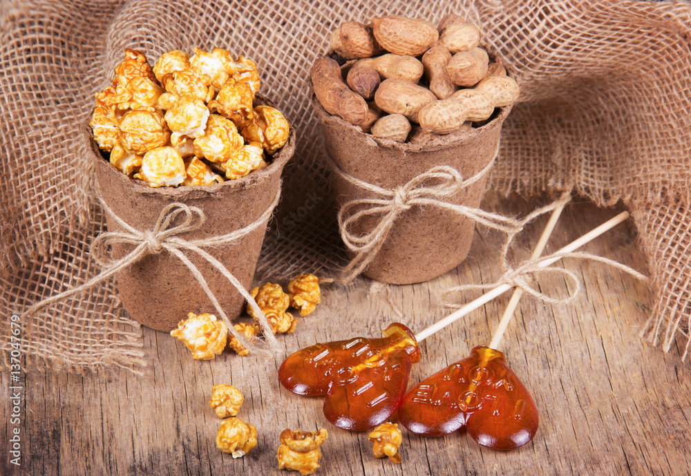Fototapeta premium Paper bucket of caramel popcorn, roasted peanuts in shell, and lollipops. Set meal for a movie. Not healthy food and snacks