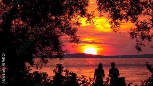 Silhouette of two people watching orange sunset over lake. Beautiful nature lifestyle travel background. Summer night in Sweden, Scandinavia. 