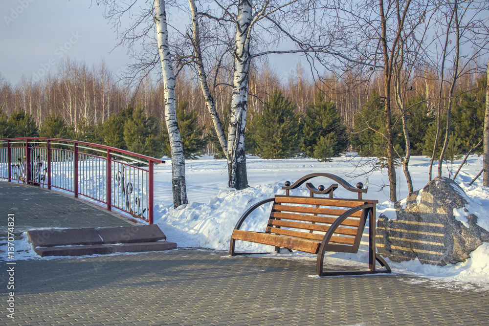 Sunny winter landscape with bench and bridge over frozen river