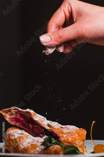 Sprinkling hot cherry pie with sugar powder. Baker finishing sweet berry dessert, free space on black background. Cooking, restaurant, bakery, pastry concept