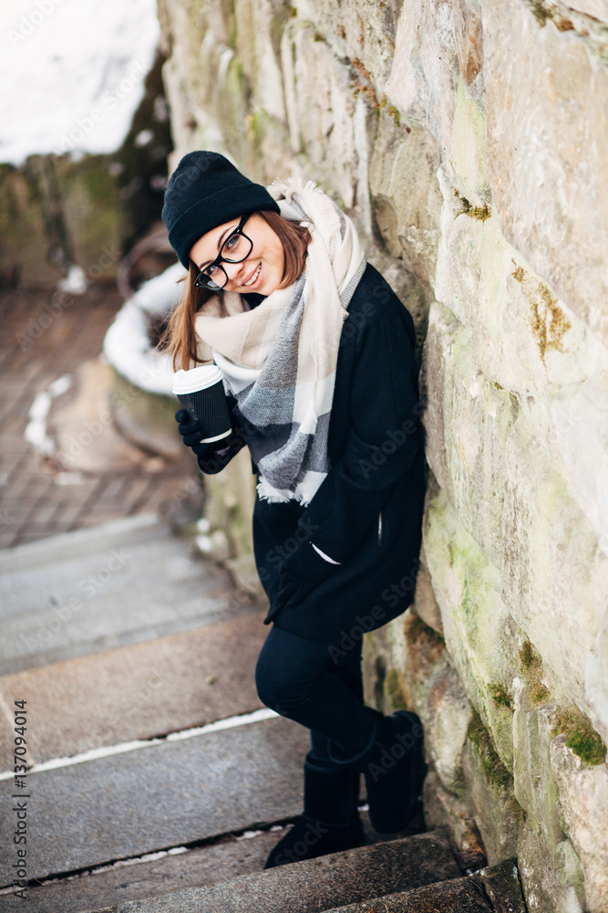 Portrait of a beautiful girl with blue eyes in black glasses in a hat and coat and gloves with scarf in the winter park holding warm coffee Portrait of a beautiful girl with blue eyes in black glasses in a hat and coat and gloves with scarf in the winter park holding warm coffee