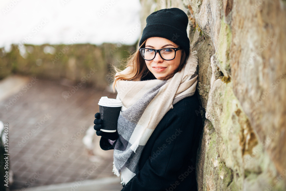 Portrait of a beautiful girl in black glasses in a hat and coat and gloves with scarf in the winter park holding warm coffee Portrait of a beautiful girl in black glasses in a hat and coat and gloves with scarf in the winter park holding warm coffee