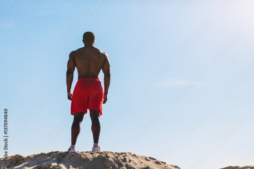 Young African American muscle man in red shorts standing on a hill on a background of blue sky in the morning. Back view