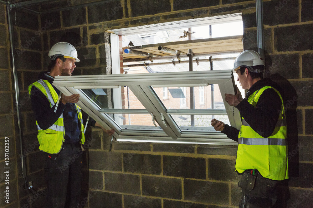 Two builders in hard hats and high visibility jacket fitting a window frame. Two builders in hard hats and high visibility jacket fitting a window frame.