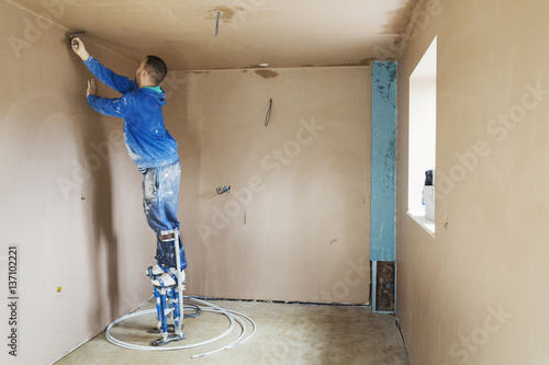 A plasterer wearing stilts smoothing fresh plaster high up on the walls of a house under construction. 