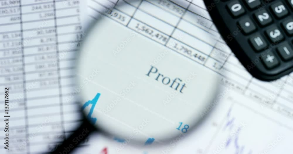 A hand with a magnifying glass passes over a financial newspaper and financial bag with a business plan. The lens magnifies the word A hand with a magnifying glass passes over a financial newspaper and financial bag with a business plan. The lens magnifies the word