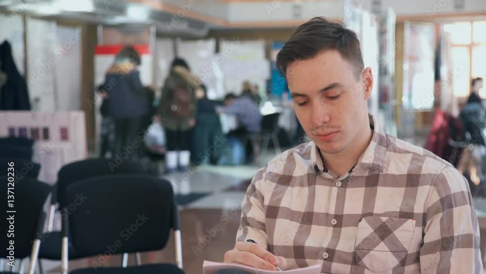 Young man sitting in a public place and making pencil writing on paper. Model with a mustache wearing a plaid shirt with a pensive look on his knee holding a notebook and record your observations. Not Young man sitting in a public place and making pencil writing on paper. Model with a mustache wearing a plaid shirt with a pensive look on his knee holding a notebook and record your observations. Not