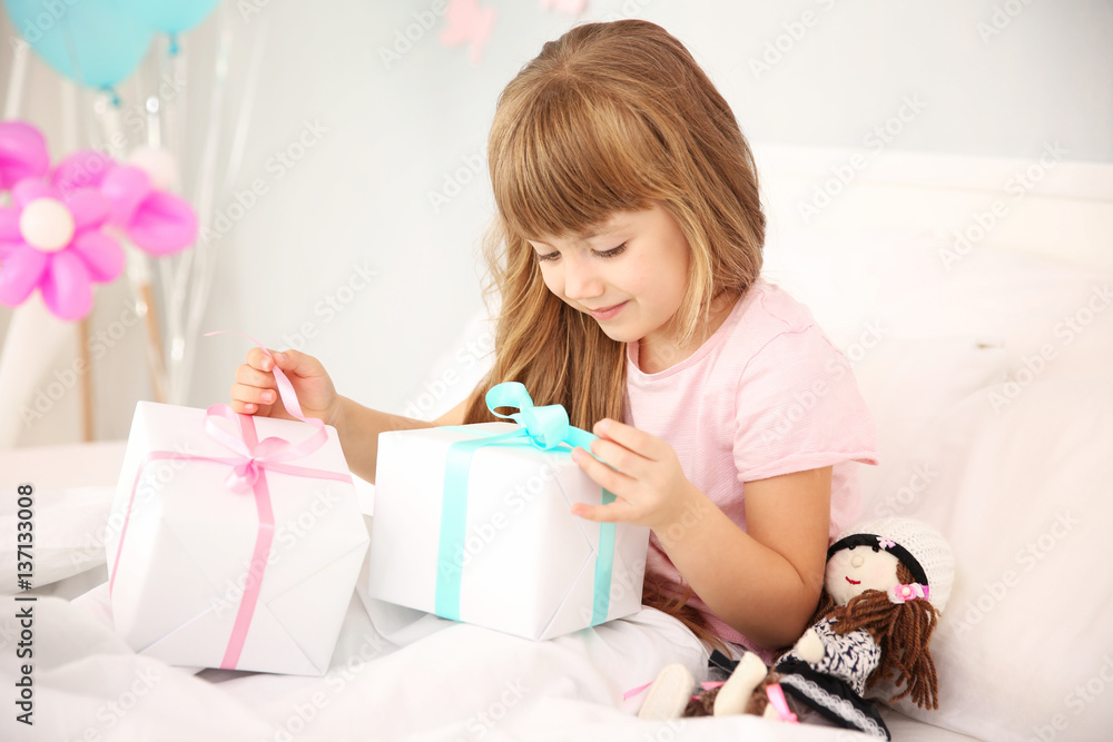 Cute birthday girl opening boxes with presents while sitting on bed at home