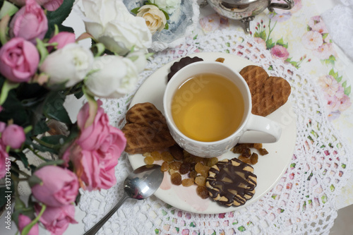 Afternoon tea time party. White cup of green light tea with cakes raisin, chocolate cookies in shabby chic elegant traditional interior on lace napkin with pink flowers. Cookie heart shape. 