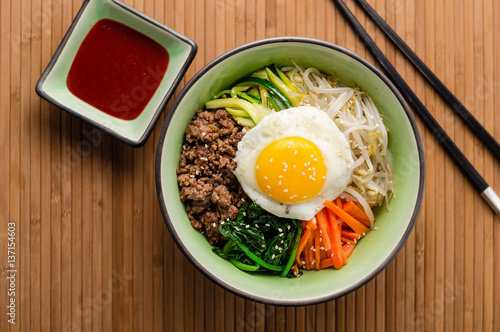 Top view of Bibimbap, a classic Korean meal. Rice is topped with seasoned vegetables, meat and a sunny side up fried egg on top. Spicy chili sauce can be added to finish off this savoury Asian dish.