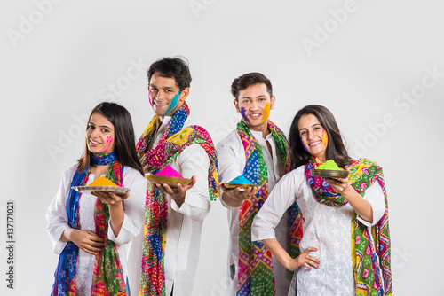 4 indian friends or 2 young couples celebrating Holi festival with sweets, bhang, pichkari and hands painted with gulal having copy space showing white board, isolated over white background