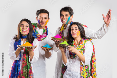 4 indian friends or 2 young couples celebrating Holi festival with sweets, bhang, pichkari and hands painted with gulal having copy space showing white board, isolated over white background