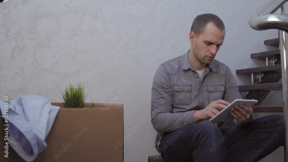 A mature man sitting on the steps of a curved staircase with lovely sunlight coming through the windows uses a digital tablet. A mature man sitting on the steps of a curved staircase with lovely sunlight coming through the windows uses a digital tablet.