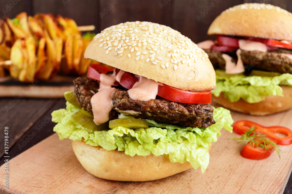 Burgers with liver cutlet, tomatoes, pickles, lettuce, spicy sauce and a soft bun with sesame seeds on a cutting board and potato slices on skewers on a dark wooden background. Close up
