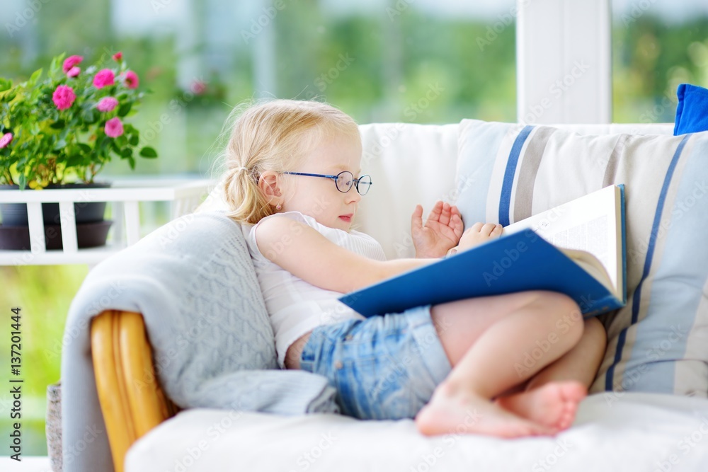 Adorable little girl wearing eyeglasses reading a book in white living room Adorable little girl wearing eyeglasses reading a book in white living room